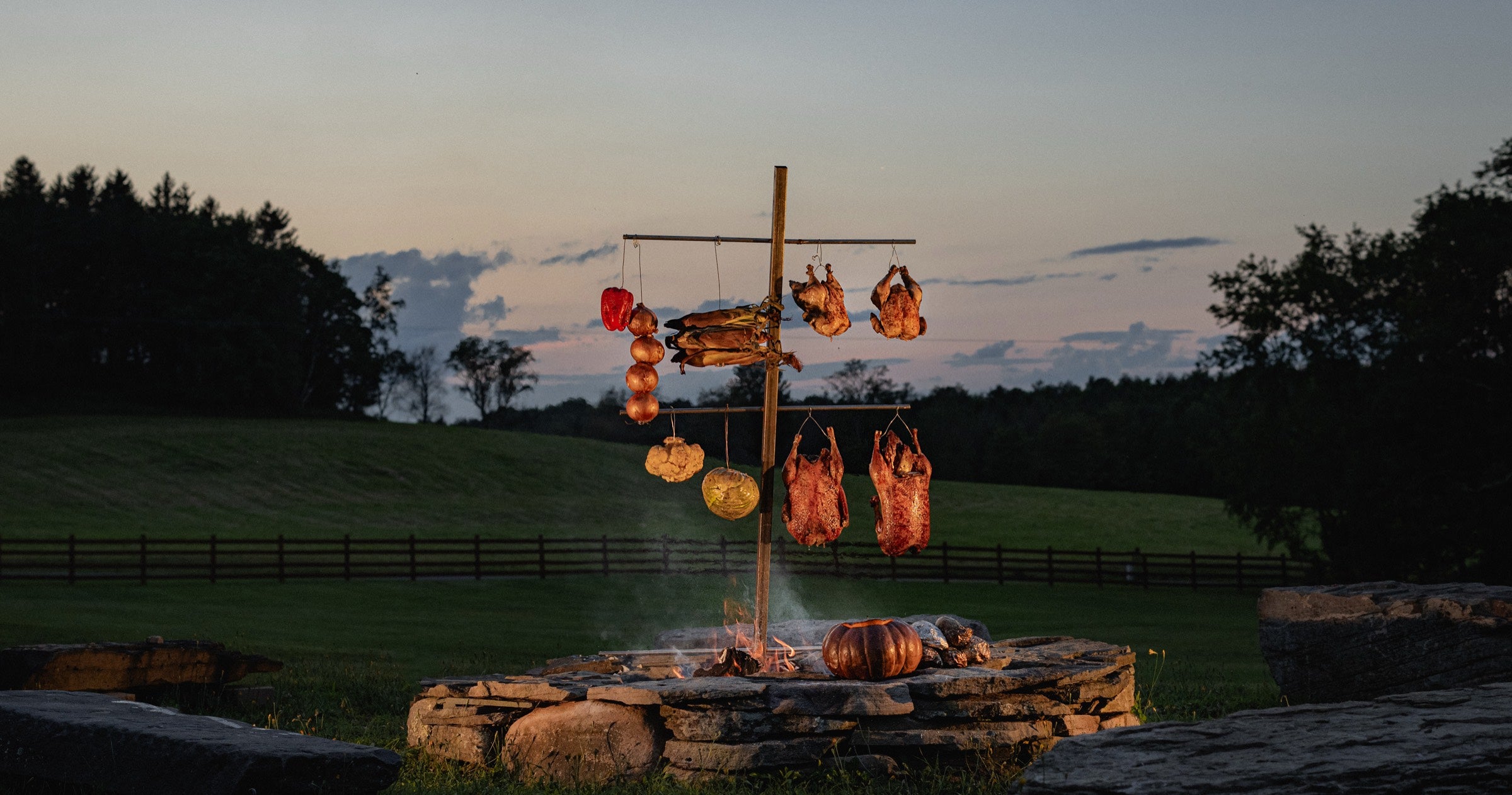 Ducks and vegetables curing on a wooden rack over the stone fire pit at dusk